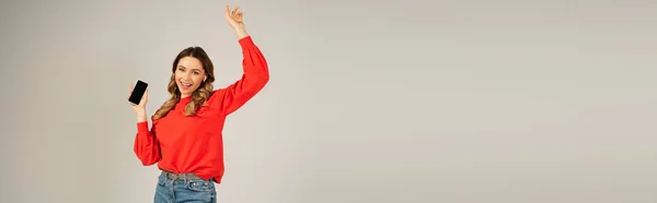 Happy woman in wireless earphones holding smartphone with blank screen isolated on grey, banner — Stock Photo