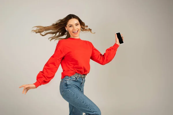 Femme excitée dans les écouteurs sans fil tenant smartphone avec écran blanc isolé sur gris — Photo de stock