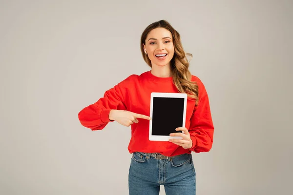 Glückliche Frau mit drahtlosen Kopfhörern, die auf ein digitales Tablet mit leerem Bildschirm zeigt, isoliert auf grau — Stockfoto