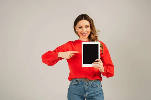 Fröhliche Frau mit drahtlosen Kopfhörern, die auf ein digitales Tablet mit leerem Bildschirm zeigt, isoliert auf grau — Stockfoto