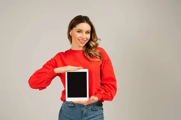 Freudige Frau mit drahtlosen Kopfhörern, die ein digitales Tablet mit leerem Bildschirm hält, isoliert auf grau — Stockfoto