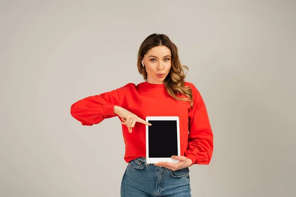 Frau mit Entengesicht in drahtlosen Kopfhörern zeigt auf digitales Tablet mit leerem Bildschirm isoliert auf grau — Stockfoto