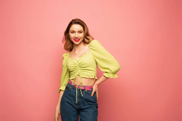 Mujer sonriente en blusa verde posando con la mano en la cadera aislada en rosa - foto de stock