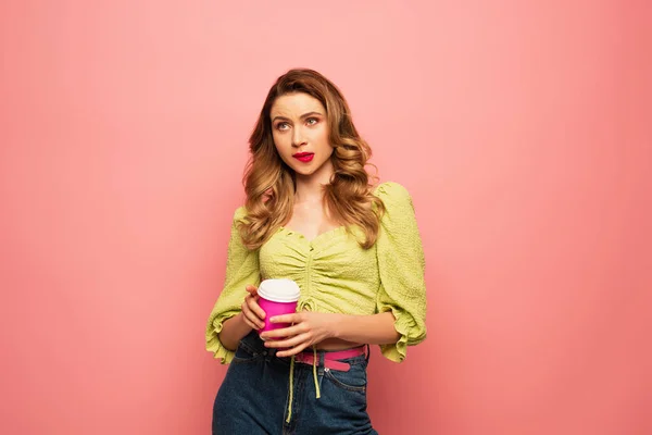 Confusa donna in camicetta verde che tiene il caffè per andare isolato su rosa — Foto stock