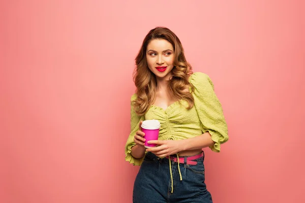 Curiosa donna in camicetta verde che tiene il caffè per andare e mordere le labbra guardando altrove isolato sul rosa — Foto stock