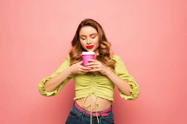 Mujer alegre en blusa verde oliendo café para ir aislado en rosa - foto de stock