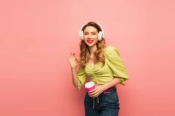 Mulher alegre em fones de ouvido sem fio segurando copo de papel e mostrando sinal ok isolado em rosa — Fotografia de Stock