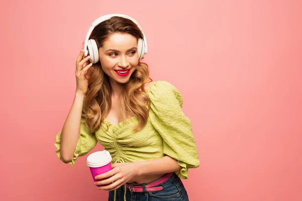 Femme joyeuse ajustant écouteurs sans fil et tenant tasse en papier isolé sur rose — Photo de stock