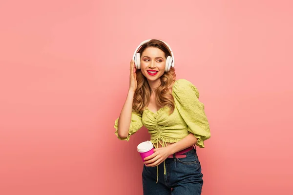 Mulher feliz ajustando fones de ouvido sem fio e segurando copo de papel em rosa — Fotografia de Stock