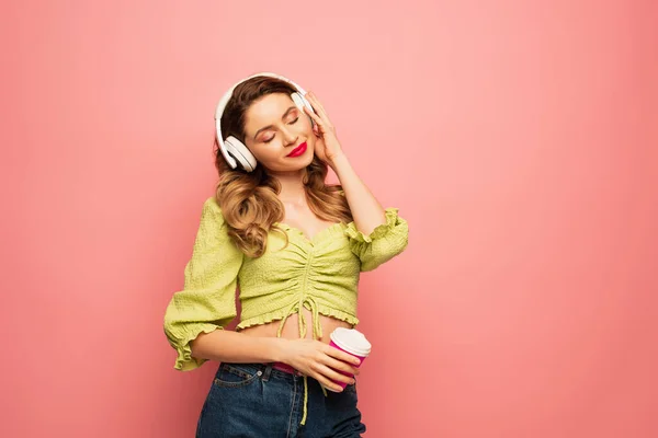 Femme souriante et bouclée ajustant écouteurs sans fil et tenant tasse en papier isolé sur rose — Photo de stock