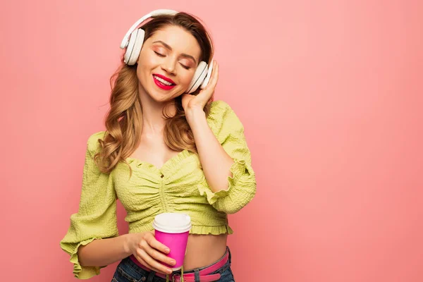 Zufriedene Frau stellt drahtlose Kopfhörer ein und hält Kaffee to go auf rosa — Stockfoto
