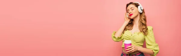 Pleased woman adjusting wireless headphones and holding coffee to go isolated on pink, banner — Stock Photo