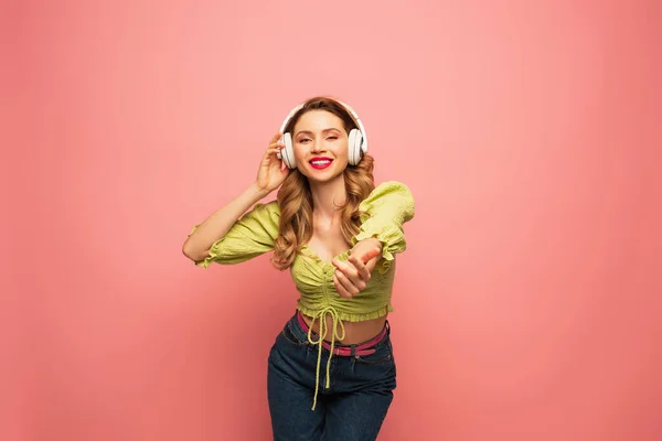 Happy woman adjusting wireless headphones and standing with outstretched hand on pink — Stock Photo