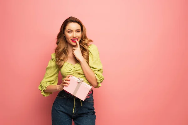 Fröhliche Frau beißt Nagel und hält umwickeltes Geschenk isoliert auf rosa — Stockfoto