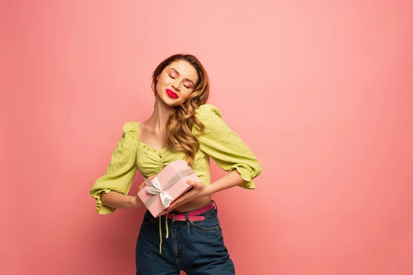 Zufriedene Frau mit geschlossenen Augen, umhülltes Geschenk isoliert auf rosa — Stockfoto
