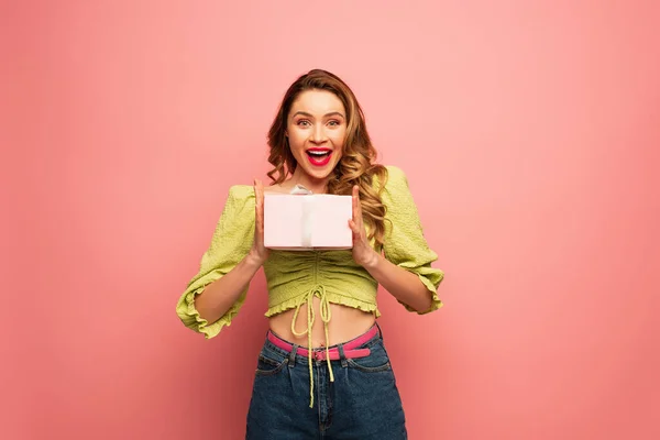 Aufgeregt und lockig Frau hält verpackten Geschenkkarton isoliert auf rosa — Stockfoto