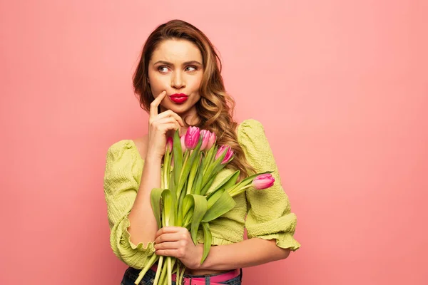 Thoughtful woman pouting lips and holding bouquet of tulips isolated on pink — Stock Photo