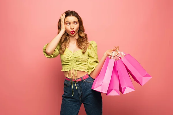 Mujer conmocionada sosteniendo bolsas de compras y pucheros labios aislados en rosa - foto de stock