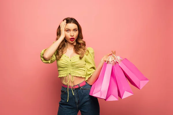 Mujer sorprendida sosteniendo bolsas de compras y mirando a la cámara aislada en rosa - foto de stock