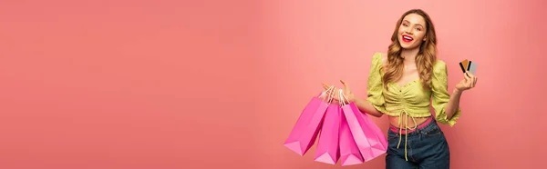 Amazed woman holding credit card and shopping bags isolated on pink, banner — Stock Photo