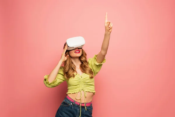 Glückliche Frau in vr Headset zeigt mit dem Finger isoliert auf rosa — Stockfoto