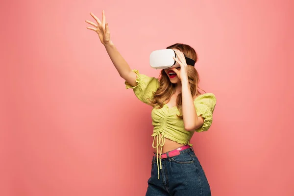 Mujer sorprendida en auriculares vr con gesto de la mano levantada aislado en rosa - foto de stock