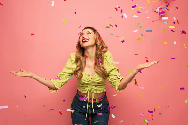 Femme excitée et bouclée avec les mains tendues près de confettis sur rose — Photo de stock