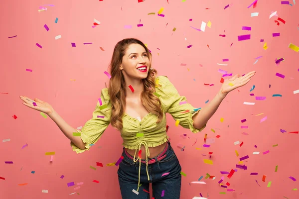 Cheerful woman with outstretched hands near confetti on pink — Stock Photo