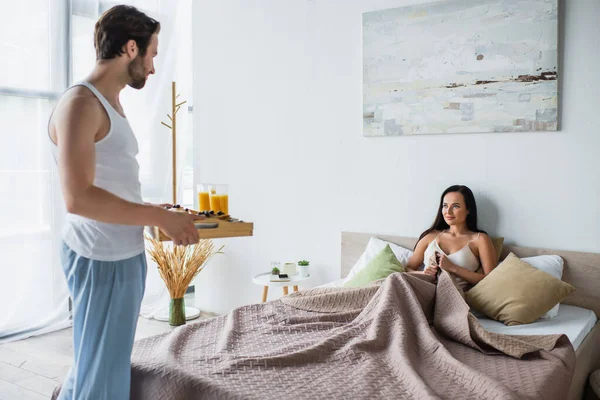 Bearded man holding breakfast tray near smiling girlfriend in bed — Stock Photo