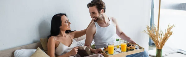 Счастливый мужчина, держащий трапезу завтрака и глядящий на веселую подругу в постели, баннер — стоковое фото