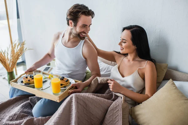 Homme heureux tenant plateau de petit déjeuner et regardant petite amie gaie au lit — Photo de stock