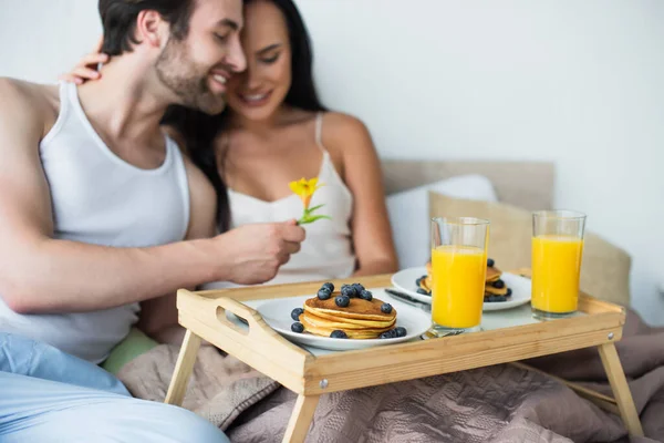 Plateau de petit déjeuner avec crêpes et jus d'orange près de couple flou et heureux au lit — Photo de stock