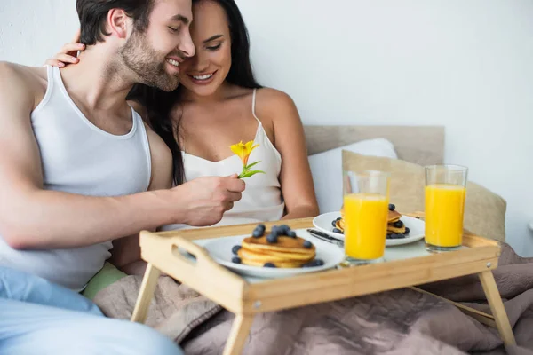 Casal feliz sorrindo e olhando para flor florescendo perto bandeja de café da manhã com panquecas e suco de laranja — Fotografia de Stock