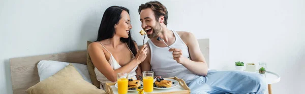 Mulher alegre alimentando namorado com panquecas saborosas na cama, banner — Fotografia de Stock