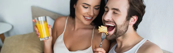 Smiling woman feeding boyfriend with tasty pancakes and holding glass of orange juice, banner — Stock Photo