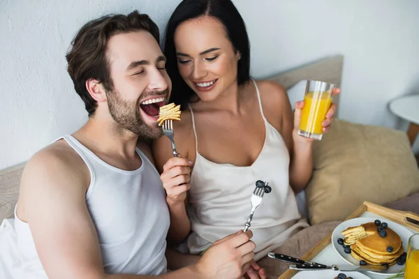 Donna sorridente che nutre il fidanzato con gustose frittelle e un bicchiere di succo d'arancia — Foto stock
