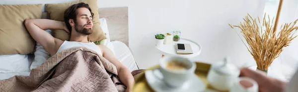 Mujer borrosa sosteniendo bandeja con taza de café cerca del hombre dormido, pancarta - foto de stock