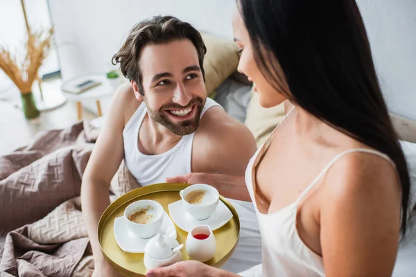 Donna sfocata e gioiosa che tiene vassoio con tazze di caffè vicino fidanzato felice a letto — Foto stock