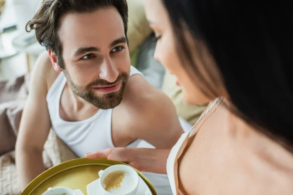 Donna sfocata che tiene vassoio con tazze di caffè vicino fidanzato barbuto a letto — Foto stock