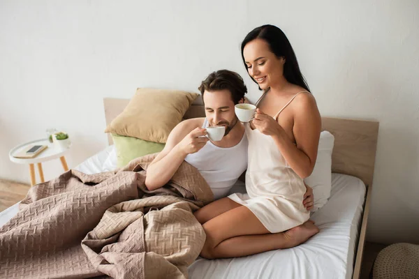 Happy couple holding cups and drinking coffee in bed — Stock Photo