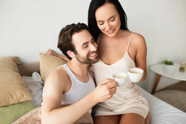 Feliz pareja tazas tintineo de capuchino en la cama - foto de stock