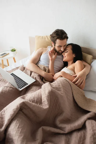 Feliz pareja acostada en la cama abrazando cerca de la computadora portátil - foto de stock