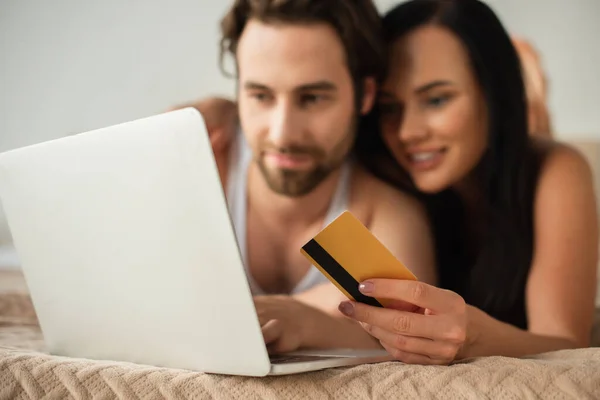 Pareja borrosa celebración de la tarjeta de crédito cerca del ordenador portátil, mientras que las compras en línea en el dormitorio - foto de stock