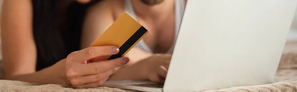 Обрезанный вид пары, держащей кредитную карту возле ноутбука во время покупок в Интернете в спальне, баннер — стоковое фото