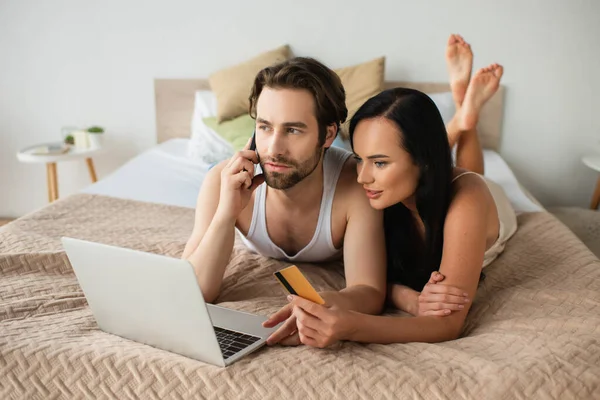 Homme tenant carte de crédit et parler sur smartphone près d'un ordinateur portable tout en faisant des achats en ligne avec petite amie — Photo de stock
