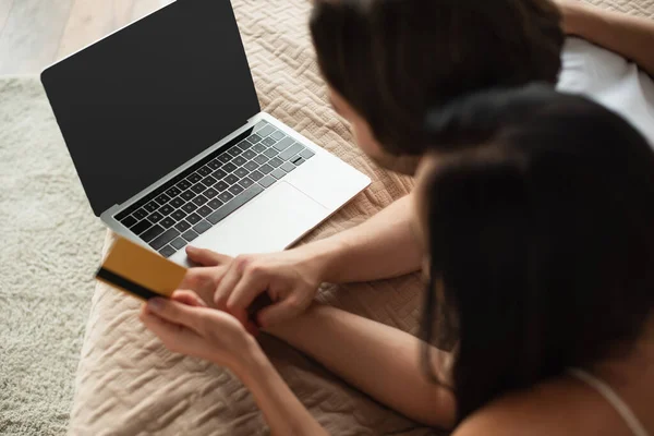 Vue grand angle du couple tenant la carte de crédit près de l'ordinateur portable avec écran blanc tout en faisant des achats en ligne dans la chambre — Photo de stock