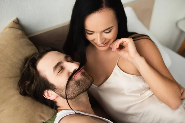 Happy woman looking at man making mustache of her brunette hair in bed — Stock Photo