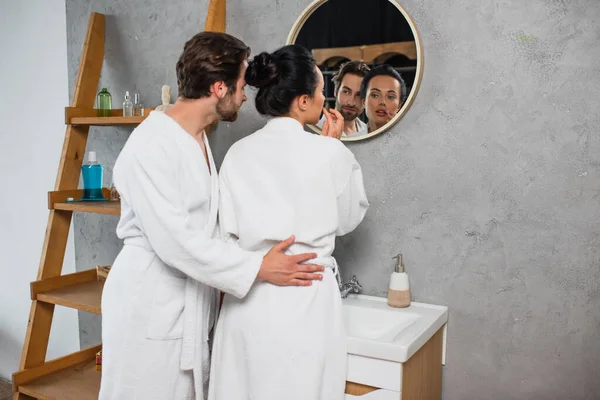 Junges Paar in weißen Bademänteln mit Blick auf Spiegel im Badezimmer — Stockfoto