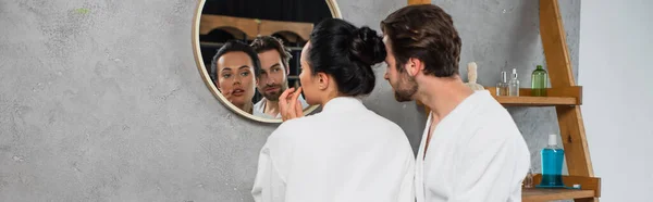 Young couple in white bathrobes looking at mirror in bathroom, banner — Stock Photo