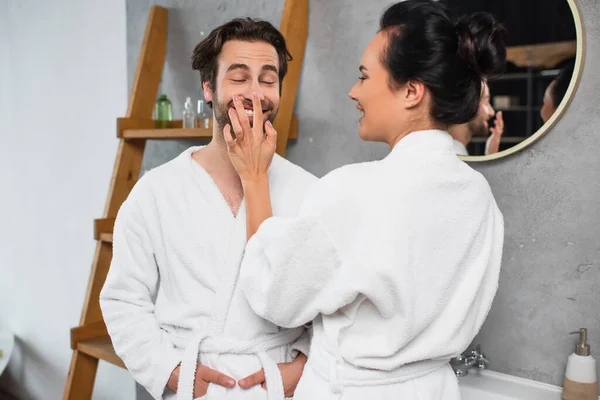 Щаслива жінка наносить крем для обличчя на ніс усміхненого хлопця в халаті — стокове фото